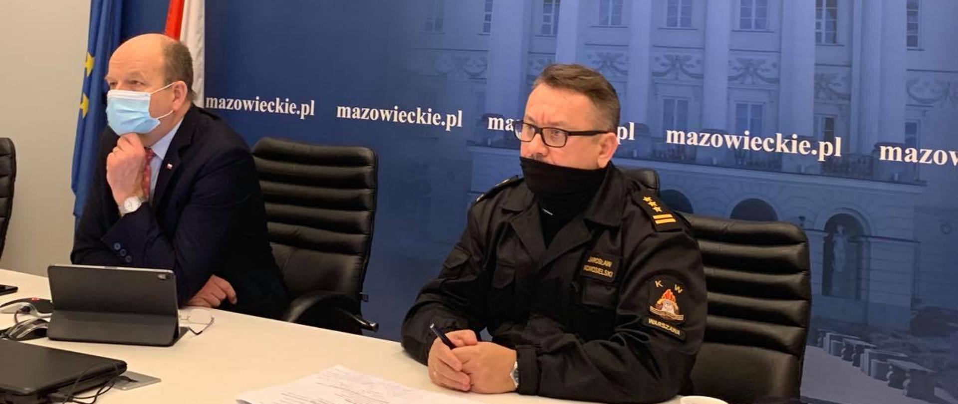 Elegancko ubrany mężczyzna (z lewej) oraz mężczyzna w mundurze ( z prawej) siedzą za stołem na tle niebieskiej ścianki z białym budynkiem