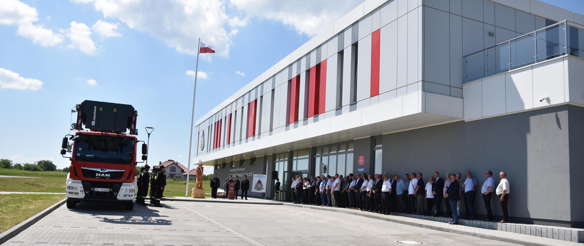 Zdjęcie przedstawia Komendanta Miejskiego Państwowej Straży Pożarnej w Rzeszowie wraz ze swoimi Zastępcami, Wojewódzkim Kapelanem Strażaków, Oficerem Prasowym Komendanta Miejskiego Państwowej Straży Pożarnej w Rzeszowie, członków Koła Emerytów i Rencistów Pożarnictwa rzeczypospolitej Polskiej przy Komendzie Miejskiej Państwowej Straży Pożarnej w Rzeszowie oraz strażaków z Jednostki Ratowniczo-Gaśniczej Nr 3 w Rzeszowie stojących na placu zewnętrznym przed strażnicą. Na wprost stoi drewniana rzeźba św. Floriana. Komendant Miejski Państwowej Straży Pożarnej w Rzeszowie przemawia przy mównicy.