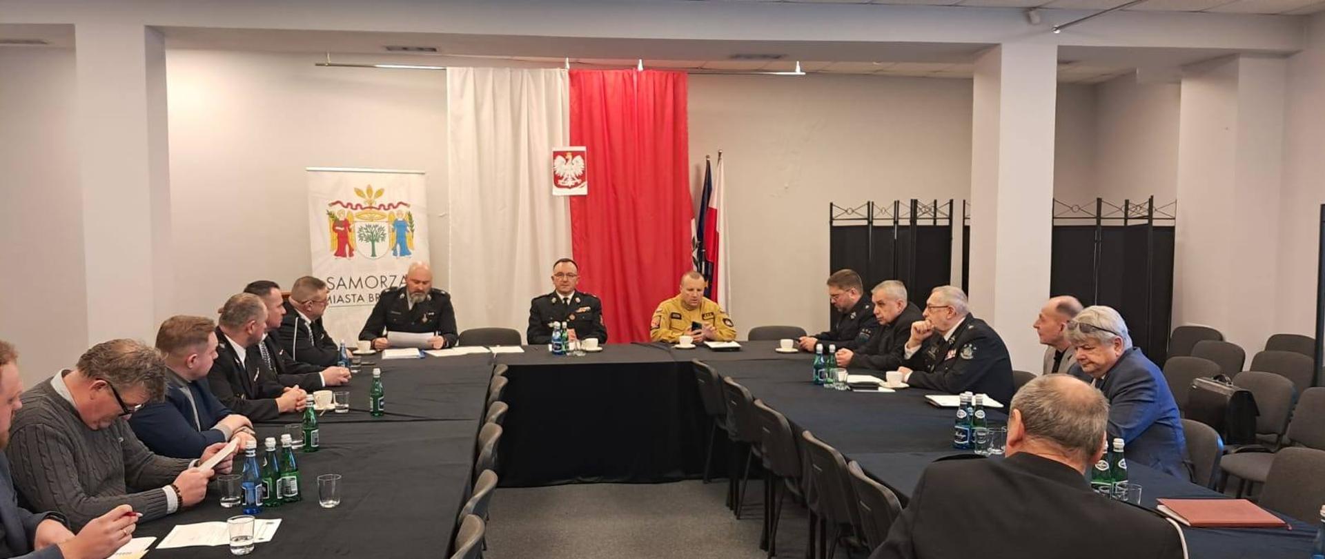 Za stołami z prawej lewej i na wprost siedzą przedstawiciele samorządu oraz strażacy. W tle w pionie flaga Polski.