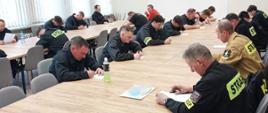 Strażacy ubrani w czarne i piaskowe ubrania koszarowe w trakcie kursu w sali szkoleniowej, siedzą przy ławkach na których leżą białe karty, ponadto w tle widać biały ekran rzutnika