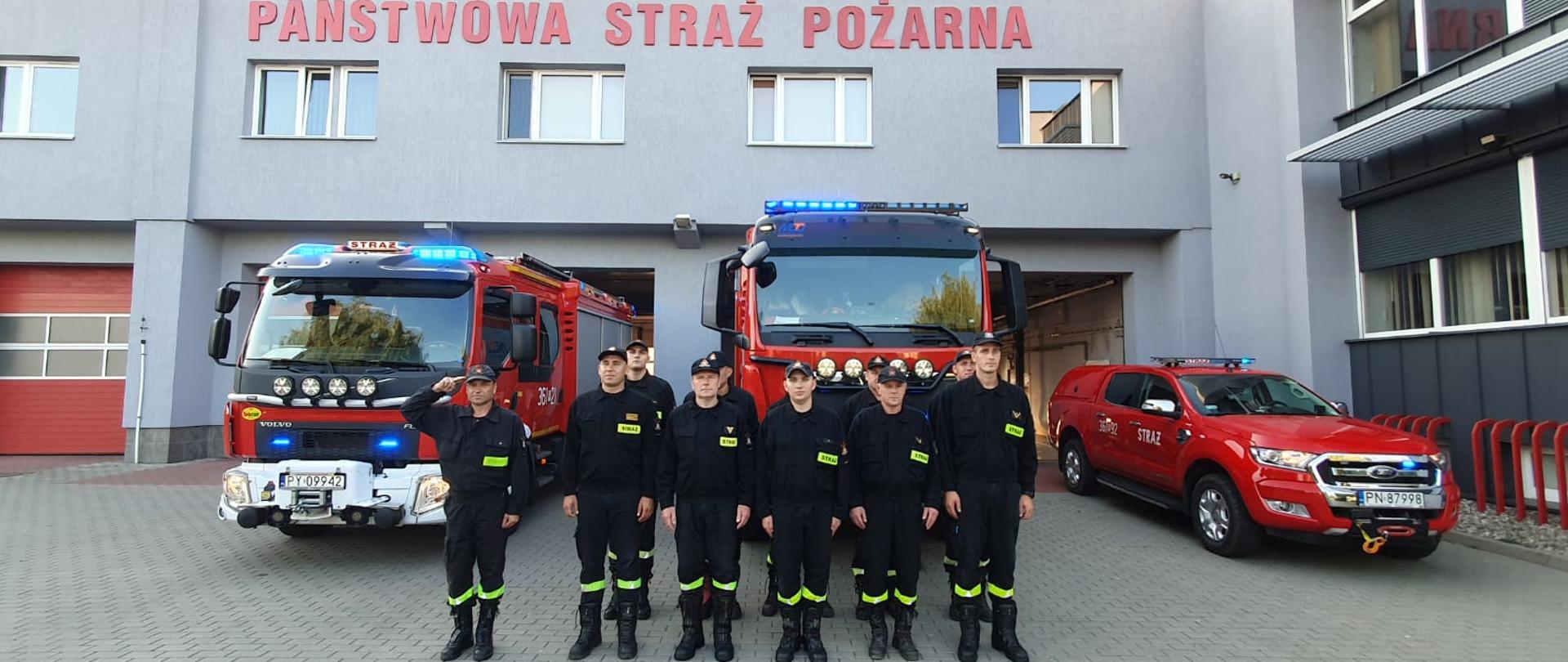 Zdjęcie przedstawia strażaków stojących przed strażnicą podczas oddania hołdu zmarłemu druhowi. Z tyłu stoją samochody strażackie na włączonych sygnałach świetlnych. 