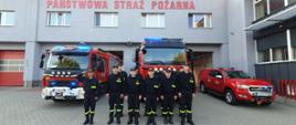 Zdjęcie przedstawia strażaków stojących przed strażnicą podczas oddania hołdu zmarłemu druhowi. Z tyłu stoją samochody strażackie na włączonych sygnałach świetlnych. 
