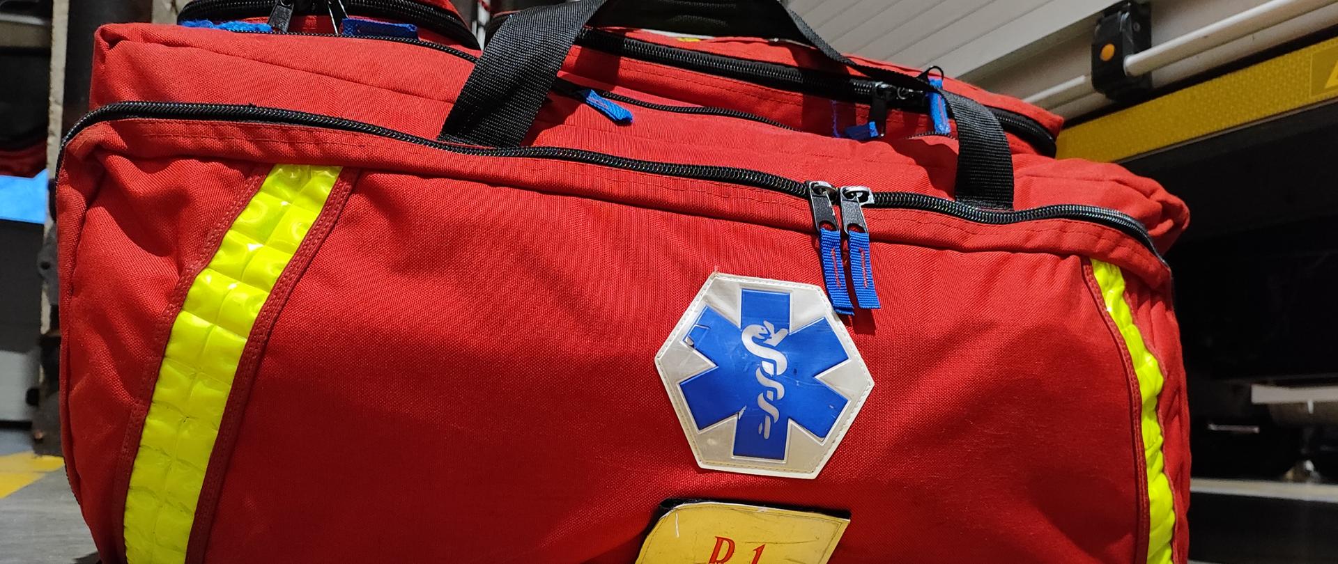 Zdjęcie przedstawiające torbę medyczną PSP-R1 na tle samochodów ratowniczo-gaśniczych