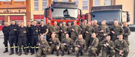 Strażacy i żołnierze wspólnie pozują do zdjęcia na placu wewnętrznym Komendy Powiatowej PSP w Ostrzeszowie. Na drugim planie pojazdy służbowe obydwu formacji. W tle budynek komendy.