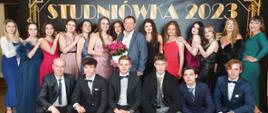 Na zdjęciu znajdują się uczniowie kl. 4C wraz z dyrektorem szkoły panem Markiem Królem Józagą i wychowawcą panem Andrzejem Mrowcą. Uczestnicy studniówki pozują do zdjęcia, znajdują się w dwóch rzędach. W pierwszym rzędzie są sami mężczyźni ubrani w białe koszule z założonymi na szyjach krawatami, bądź muszkami. Wszyscy w pierwszym rzędzie kucają. W drugim rzędzie znajdują się same dziewczyny ubrane w wieczorowe suknie. Po środku w grupie uczennic stoi wychowawca klasy. Wychowawca ubrany jest w białą koszulę i szarą marynarkę, w prawej ręce trzyma doniczkę z czerwonymi różyczkami. Z tyłu w tle znajduje się ścianka w kolorze czarnym ze złotym napisem: "Studniówka 2023" i również złotym ornamentem w kształcie ramki. Uczestnicy pozując do zdjęcia stoją na drewnianej podłodze.