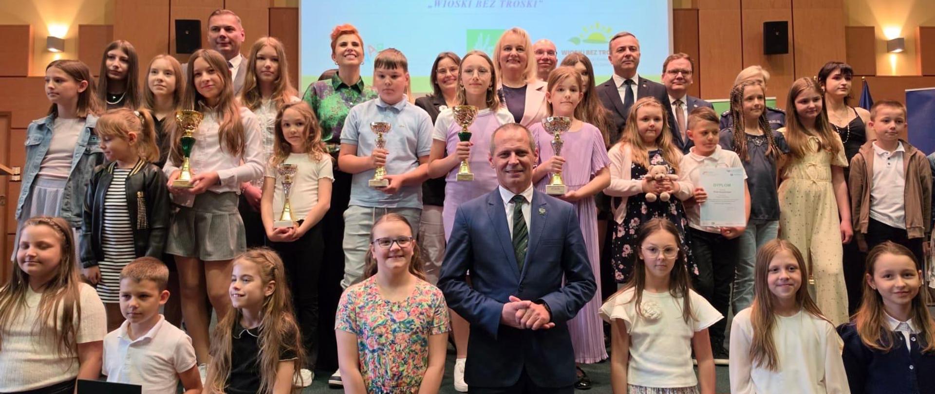 Duża grupa dzieci i dorosłych stoi na scenie. Uczestnicy są ustawieni w rzędach, a niektórzy z nich trzymają w dłoniach puchary i dyplomy. Nad grupą wisi duży ekran z napisami informującymi o finale konkursów KRUS.