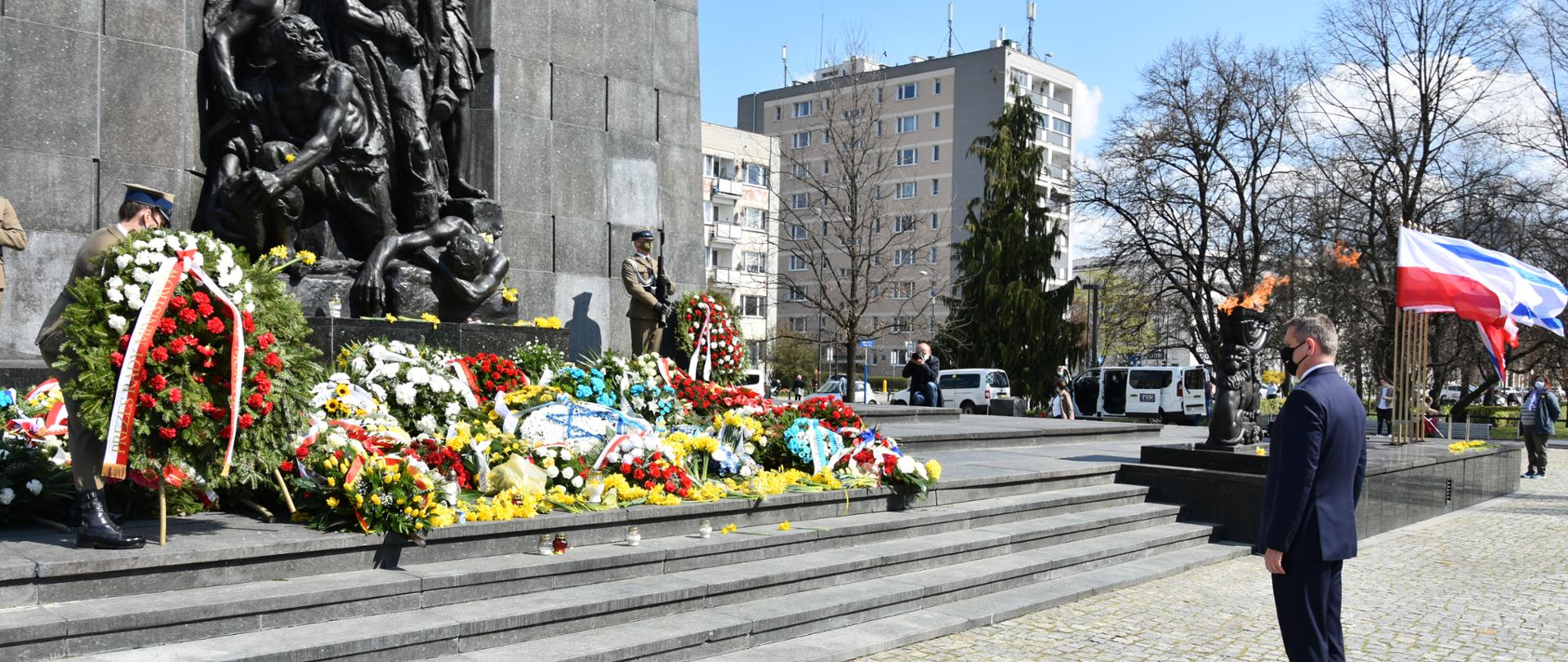 Na zdjęciu znajduje się pomnik bohaterów getta warszawskiego z kwiatami, przed pomnikiem stoi mężczyzna