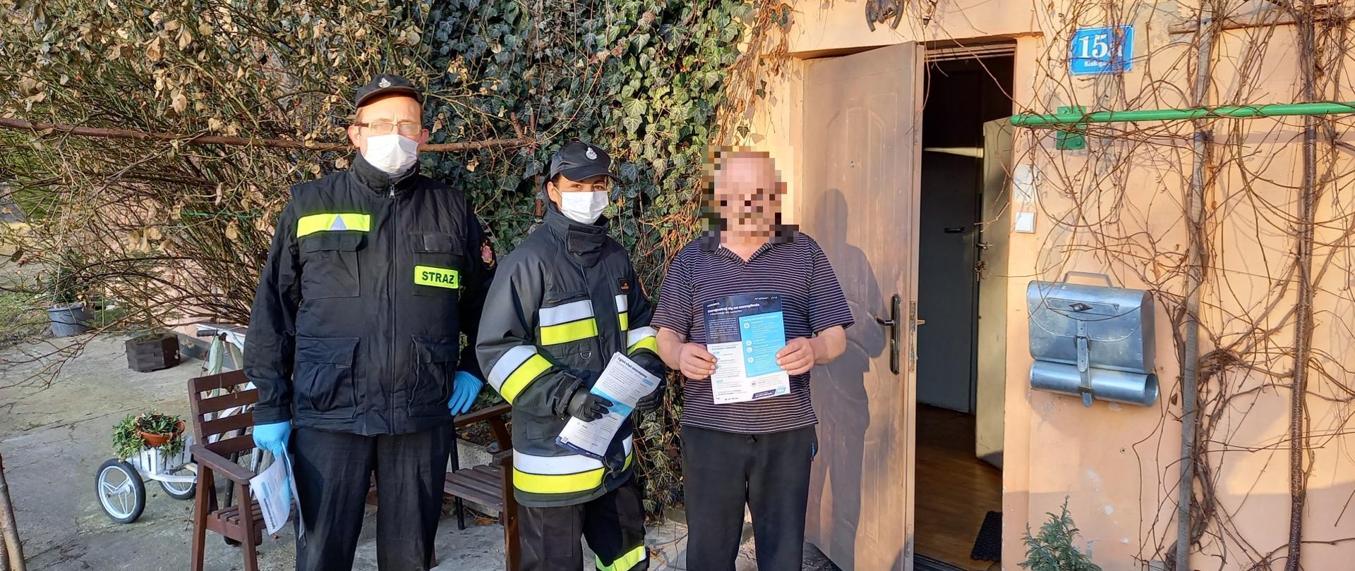 Zdjęcie przedstawia strażaków przekazujących ulotkę z informacjami dotyczącymi szczepień. Obok strażaków stoi mężczyzna trzymający przekazaną ulotkę.
