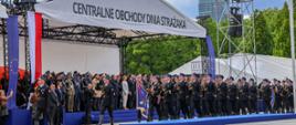 Defilada podczas obchodów Dnia Strażaka: Na zdjęciu widoczna jest scena główna z zadaszeniem oznaczonym napisem „Centralne Obchody Dnia Strażaka”. Przed sceną maszeruje kolumna funkcjonariuszy Państwowej Straży Pożarnej w galowych mundurach. W tle, na scenie, znajdują się zaproszeni goście, przedstawiciele służb i instytucji, w tym osoby w mundurach z odznaczeniami. Po bokach widać flagi Polski i Unii Europejskiej.