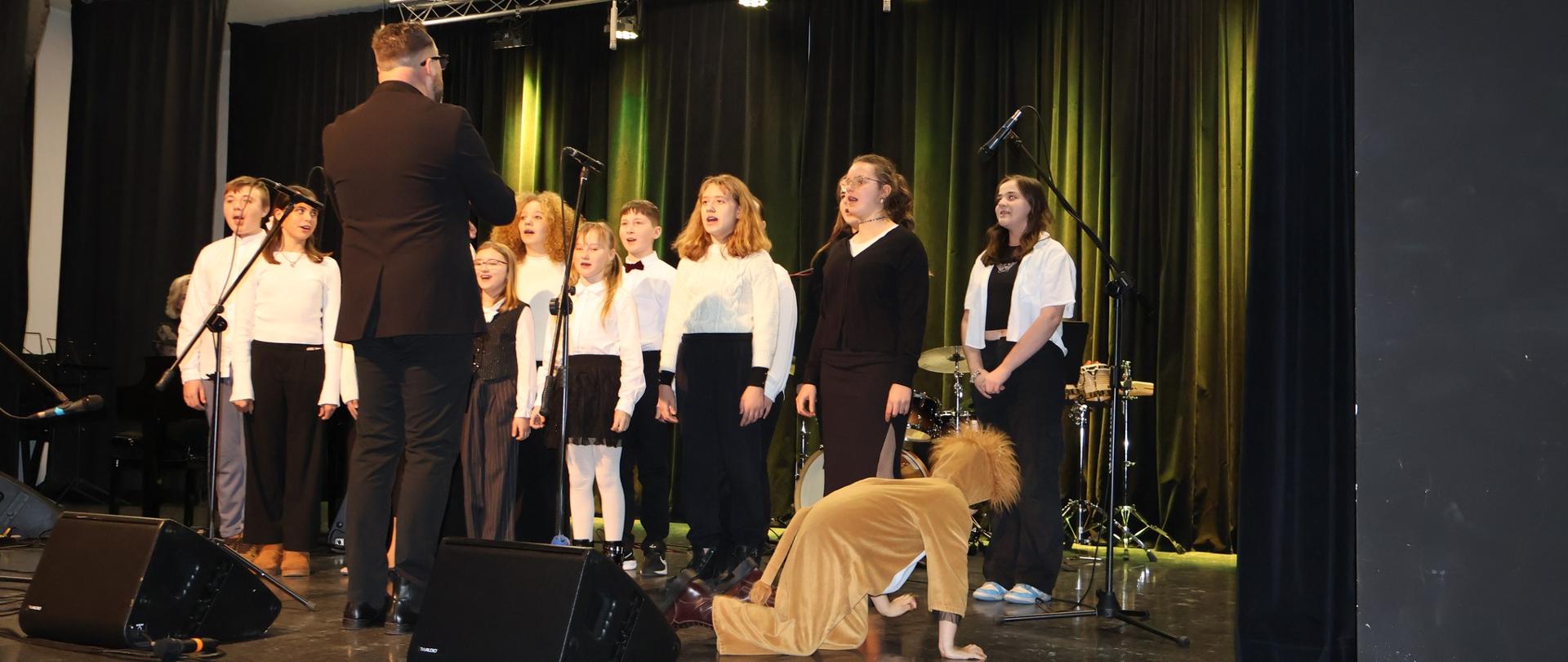 Na scenie grupa uczniów nieco zasłonięta stojącym tyłem dyrygentem. Przed chórem w przebraniu lwa uczeń.