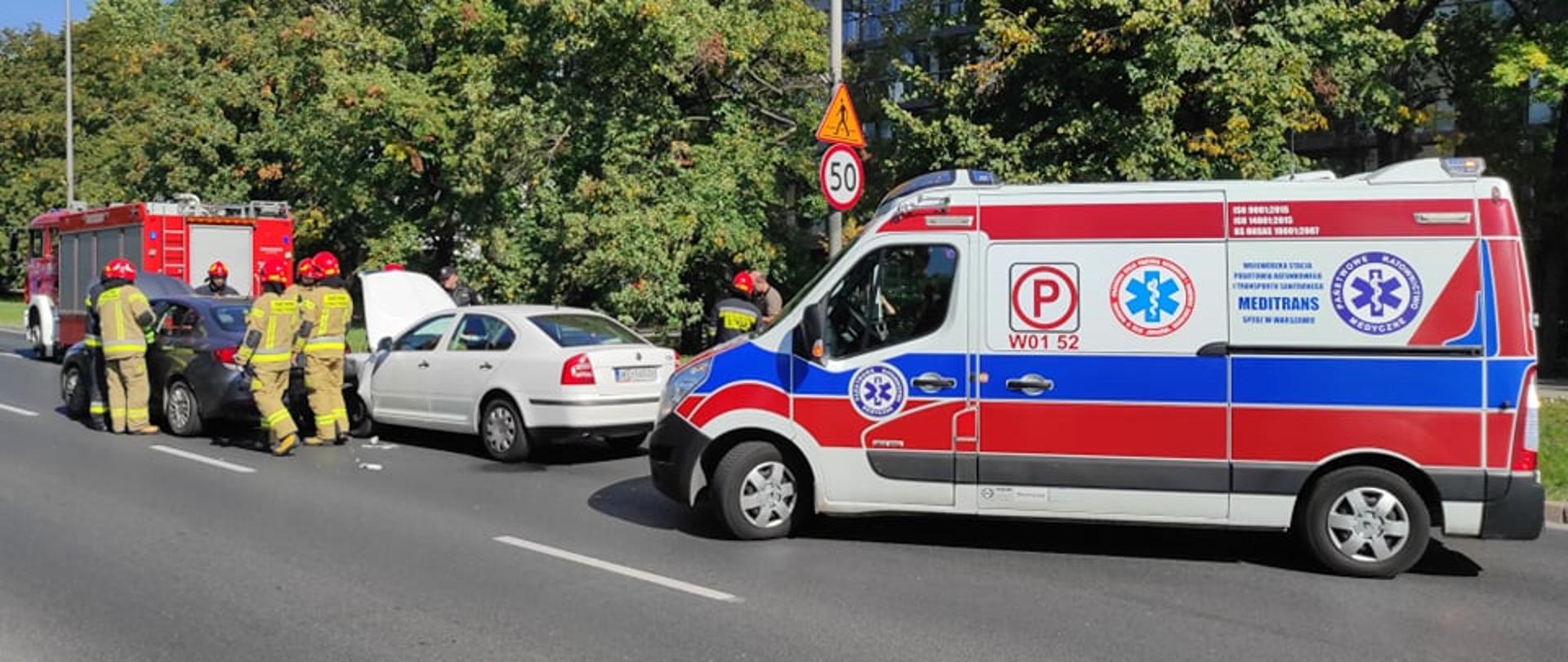 Na zdjęciu widoczna jest karetka pogotowia dalej są dwa pojazdy które brały udział w wypadku, pracujący strażacy, w tle samochód straży pożarnej.