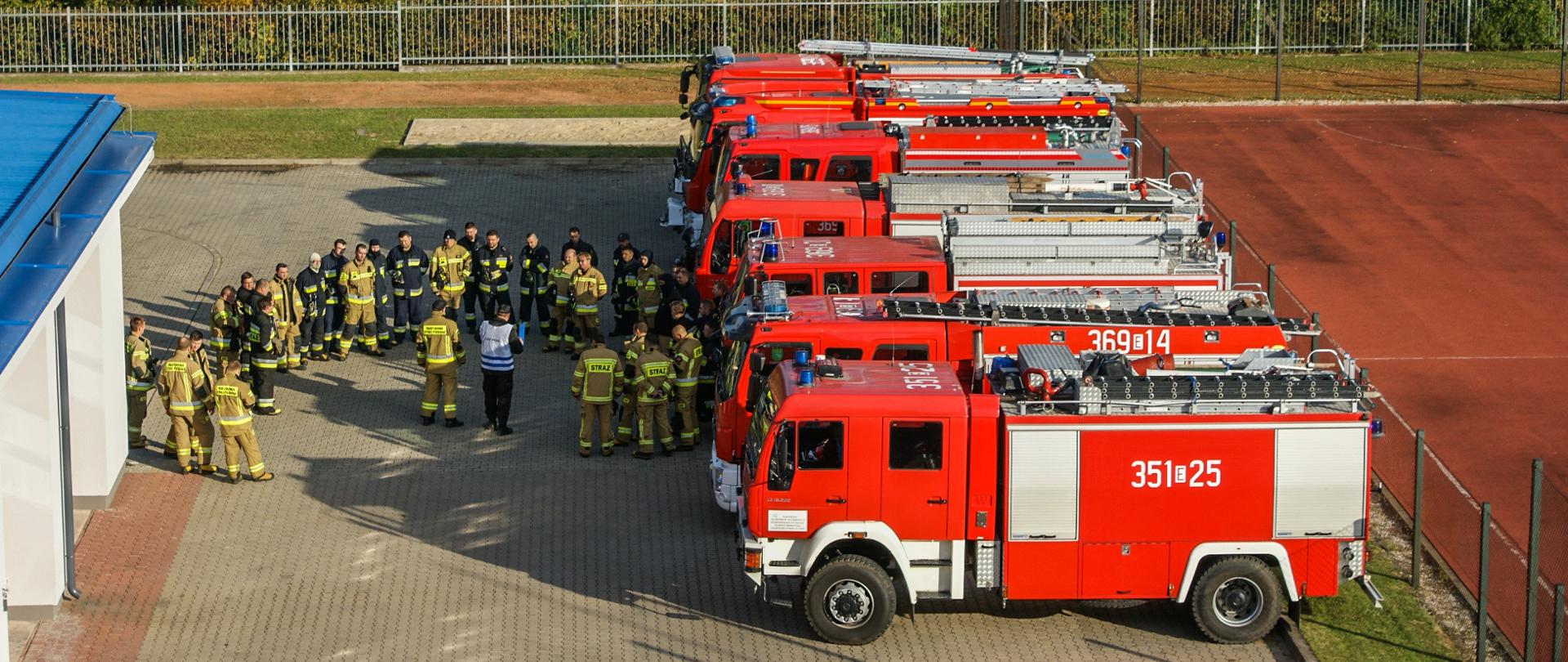 Odprawa przed ćwiczeniami na placu komendy, pojazdy ustawione na parkingu, a uczestnicy ćwiczeń są ustawieni przed nimi. 