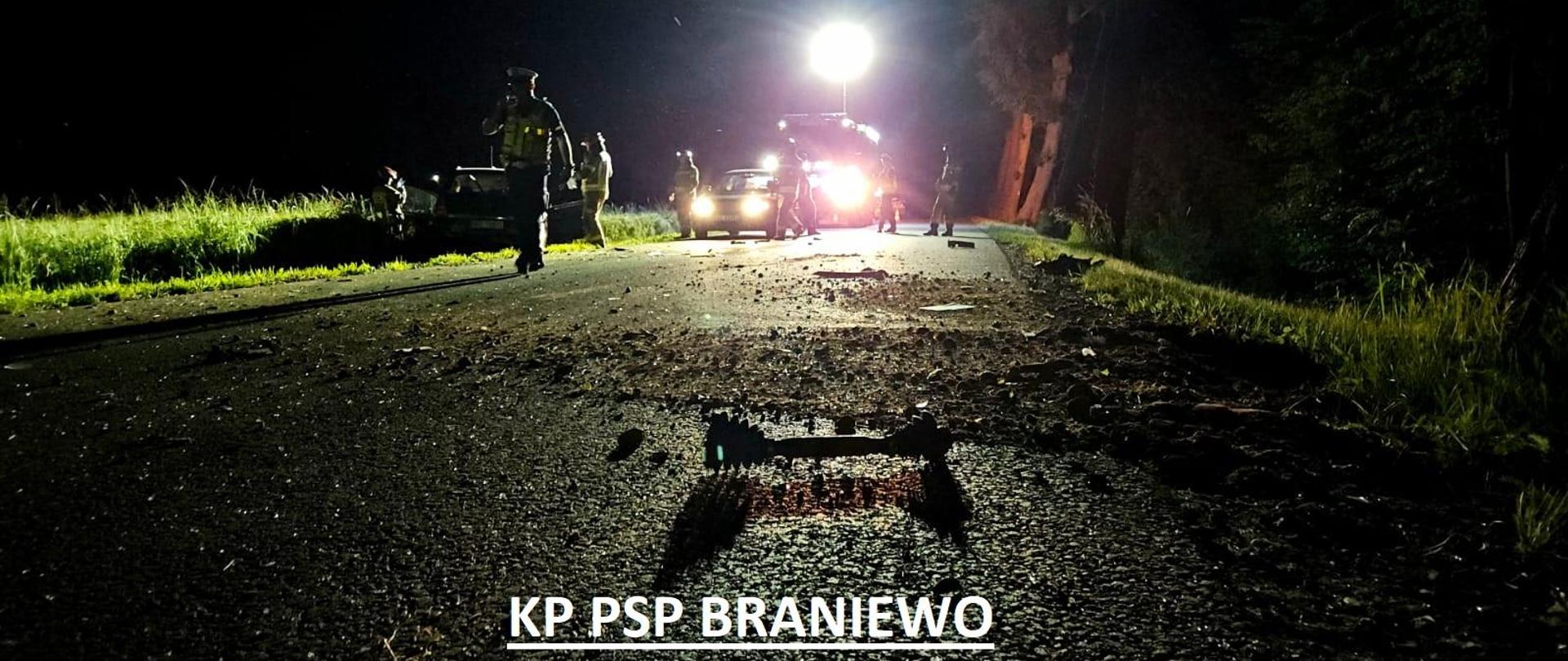 Zdjęcie zrobione nocą. W oddali samochód strażacki oświetla teren akcji. Na drodze strażacy. Poza droga auto osobowe.