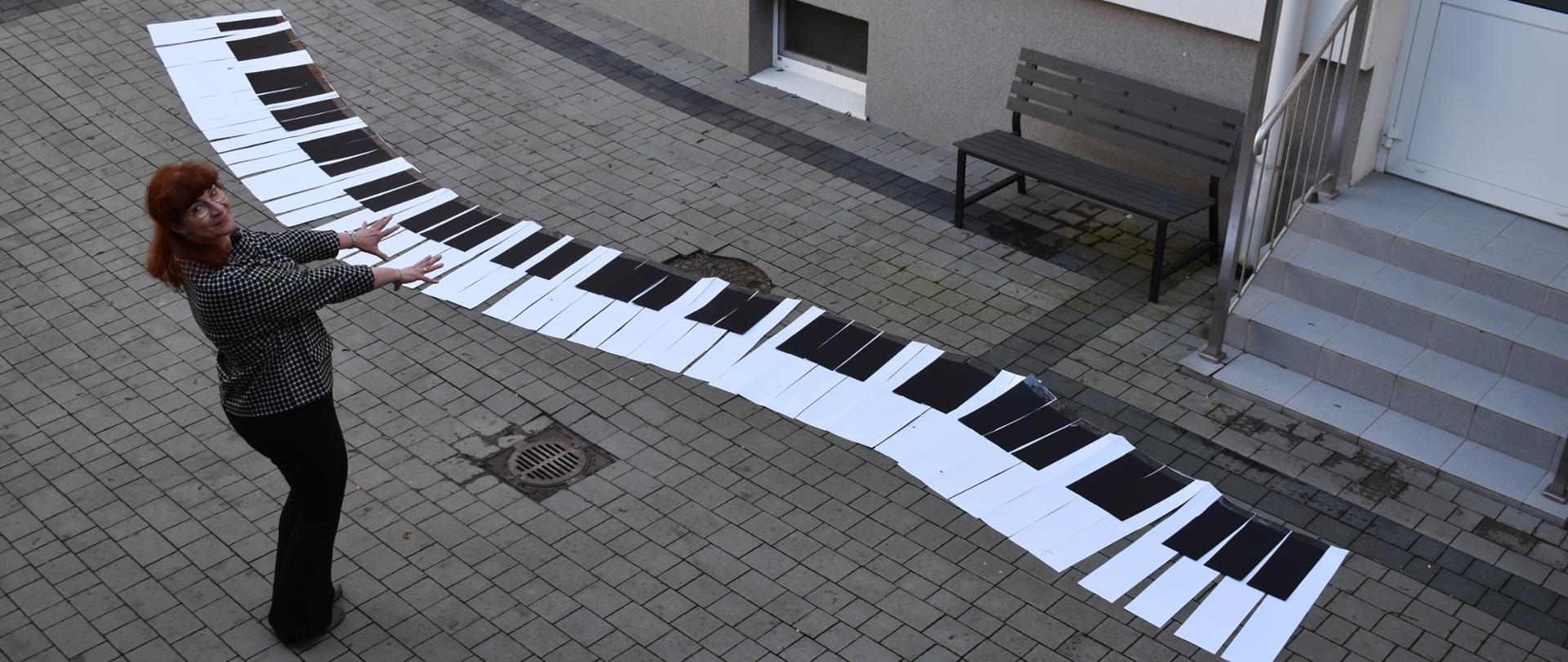 na zdjęciu widnieje dyrektor PSM I st. w Jaśle, Renata Osikowicz-Wyroba, która na dziedzińcu szkoły pozuje w charakterze pianisty, który symbolicznie gra na kilkunastometrowej klawiaturze wykonanej z kartonu. Kolorystyka zdjęcia jest biało-szaro-czarna. 