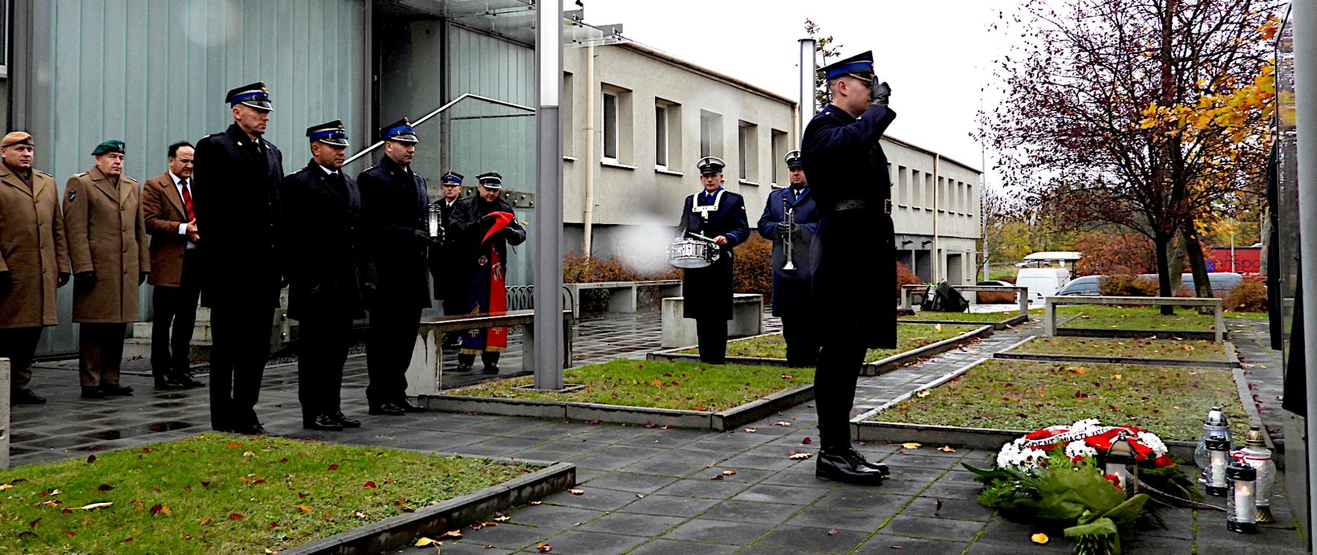 Na zdjęciu widać grupę osób biorących udział w uroczystości przed budynkiem Komendy wojewódzkiej państwowej straży pożarnej. Na szklanej fasadzie budynku widnieje napis „PAŃSTWOWA STRAŻ POŻARNA – Komenda Wojewódzka PSP w Katowicach”. Na pierwszym planie po prawej stronie stoi funkcjonariusz w granatowym mundurze wyjściowym, w rogatywce z daszkiem, salutujący w kierunku tablicy pamiątkowej. Przed nim, na ziemi, leżą wieńce i znicze. Obok niego stoją inni mundurowi zastępcy komendanta wojewódzkiego mł. bryg. Zbigniew Dyk i st. bryg. Arkadiusz Krzemiński oraz kilka osób w cywilnych ubraniach i płaszczach, ułożonych w szeregu w postawie wyprostowanej. Tło stanowi nowoczesny budynek straży oraz kilka drzew z żółtymi liśćmi – widać, że to jesień. Chodnik i trawa są mokre od deszczu.