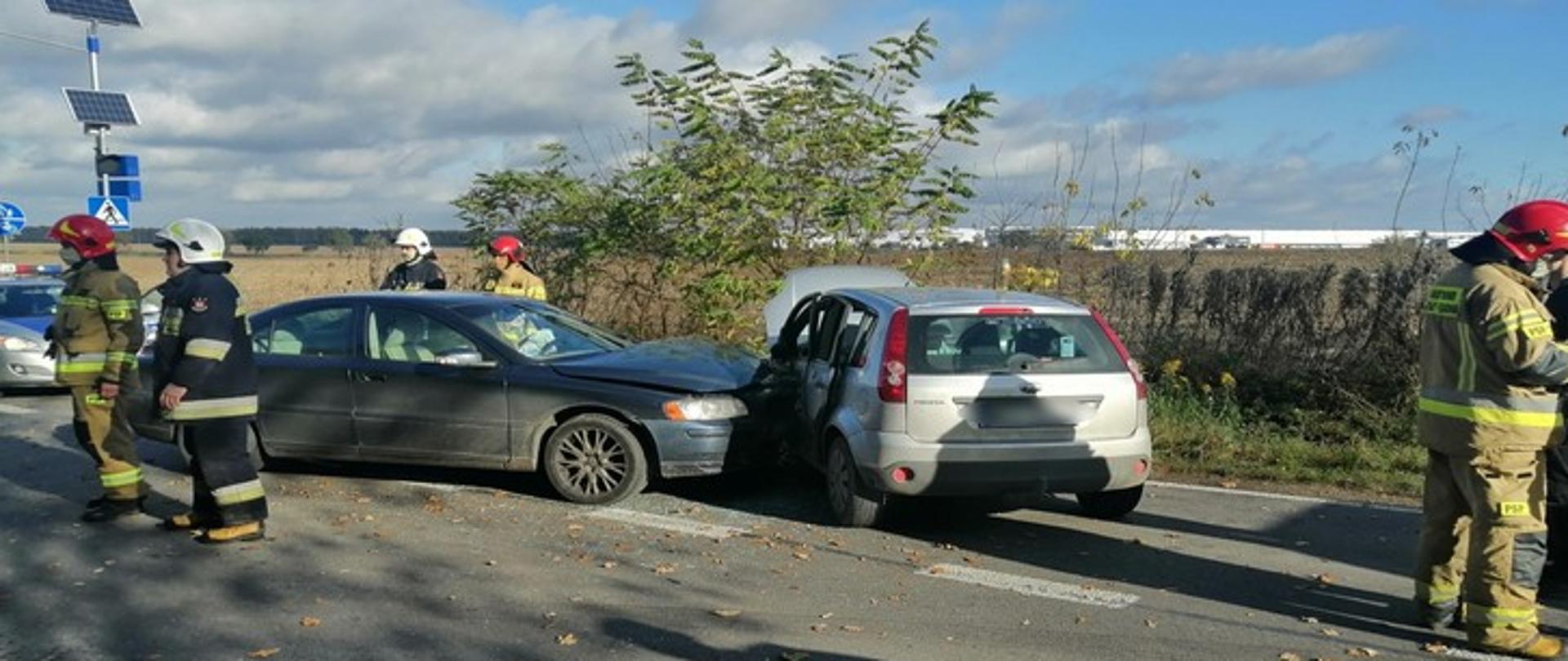 Zdjęcie przedstawia dwa samochody osobowe, które uległy wypadkowi. Pojazdy znajdują się na jezdni, a wokół nich widać strażaków ratowników.