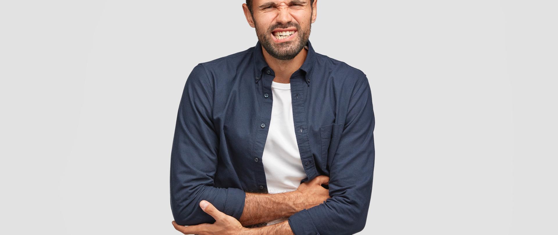 Atrakcyjny, młody brodaty mężczyzna w granatowej koszuli i białym T-shircie cierpi na okropny ból brzucha. Krzyżuje ręce i zaciska zęby z bólu brzucha.