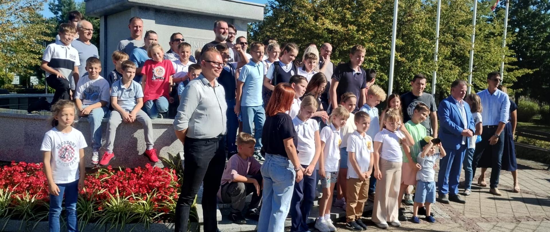 1. Na zdjęciu widać dużą grupę dzieci i dorosłych (uczestnicy turnieju szachowego) pozujących wspólnie przed pomnikiem Kazimierza Wielkiego w Kowalu, w słoneczny dzień. Część osób stoi, część siedzi na murku, a z przodu stoją dzieci i kilku dorosłych. W tle rosną drzewa i stoją maszty z flagami. Przed pomnikiem znajdują się czerwone kwiaty i zielone rośliny. Atmosfera jest radosna i pamiątkowa.