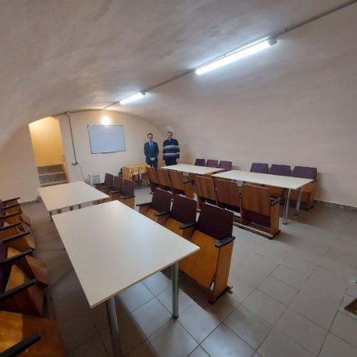 Schron, w którym znajdują się 4 stoły z krzesłami oraz tablica. Na końcu schronu stoi dwóch mężczyzn