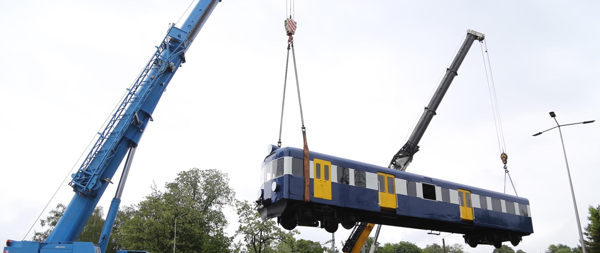Na zdjęciu widoczny wagon kolejowy wiszący w powietrzu, który jest podtrzymywany na obu końcach przez zamocowane łańcuchy widocznych w tle samochodów specjalnych typu dźwig samojezdny