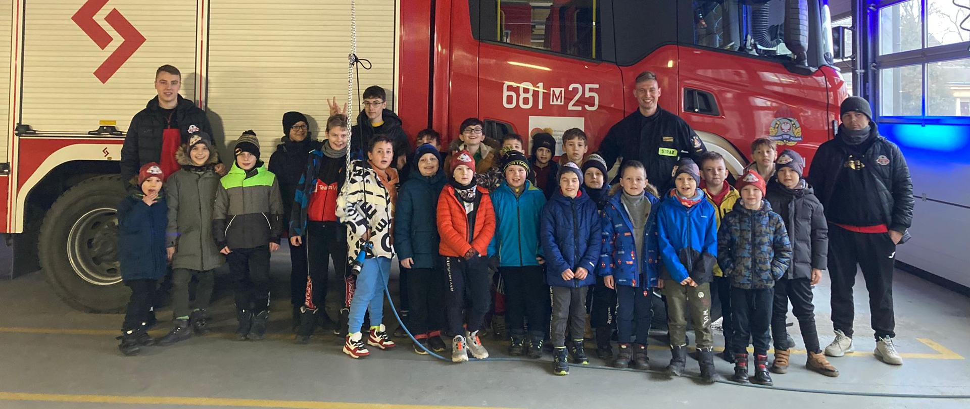 Zdjęcie przedstawia grupę dzieci stojącą na tle samochodu strażackiego. Pośród najmłodszych stroją strażacy. Auto ustawione jest w garażu. Wraz z dziećmi stoją ich opiekunowie. Wszyscy mają uśmiechnięte twarze.