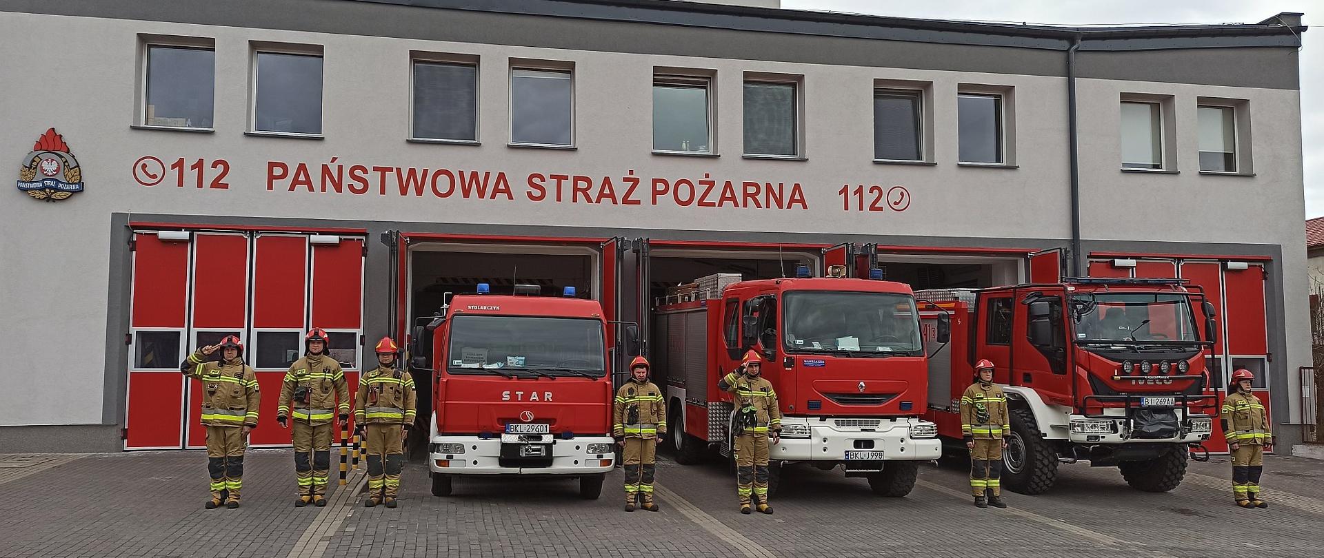 Na zdjęciu widok strażnicy i garaży. Przed garażami samochody ratowniczo-gaśnicze oraz strażacy oddający hołd tragicznie zmarłej funkcjonariuszce służby więziennej.