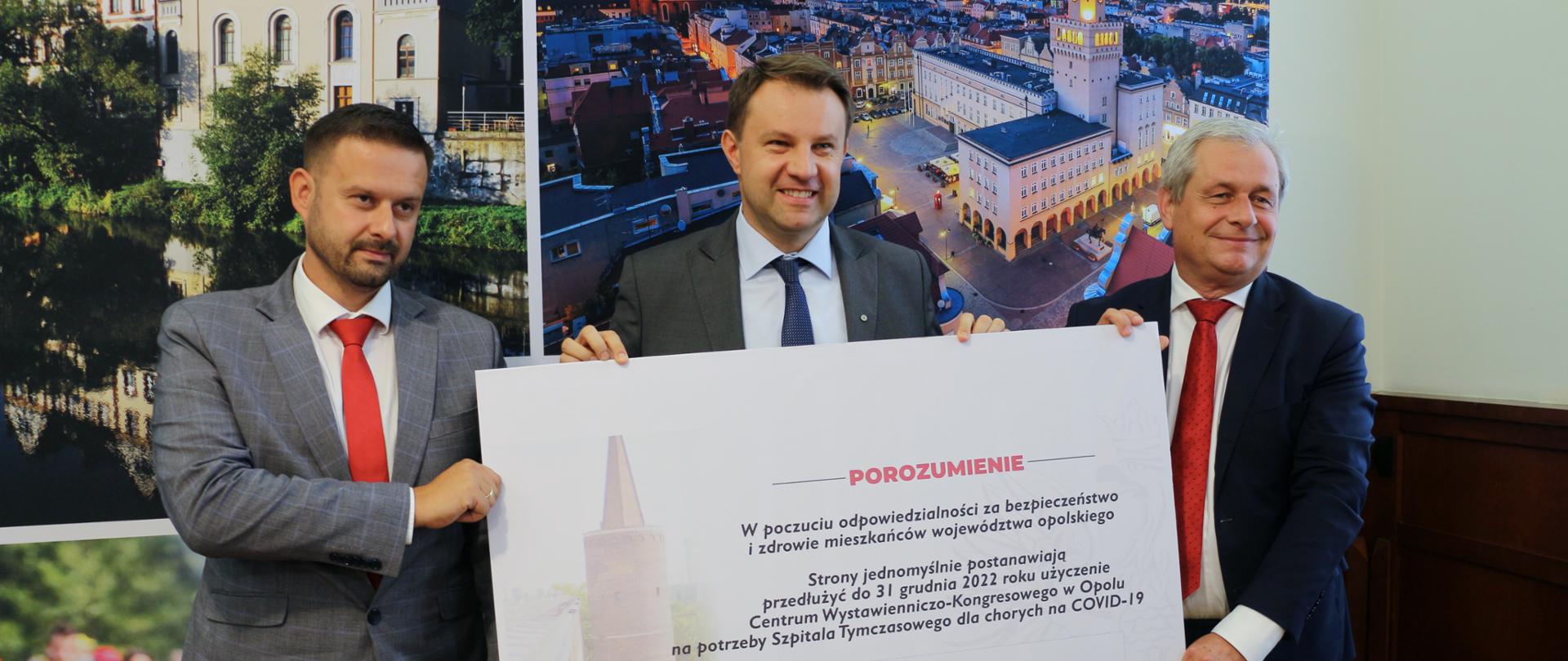 Na zdjęciu trzech mężczyzn, ubranych w garnitury. Stoją obok siebie, trzymają dużego formatu czek z informacją o zawartym porozumieniu. W tle ścianka ze zdjęciami Opola