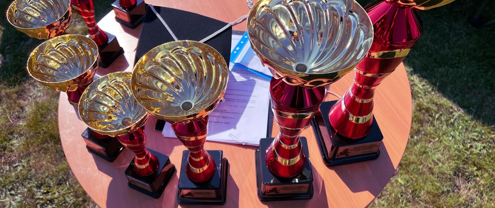 Na drewnianym stoliku w kształcie koła stoi 8 pucharów w kolorze czerwono-złotym.