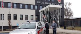 Strażak w czarnym mundurze przed budynkiem urzędu gminy, przekazuje ulotki mężczyźnie w szarej marynarce i czarnych spodniach. Obok strażaka samochód strażacki koloru srebrnego.