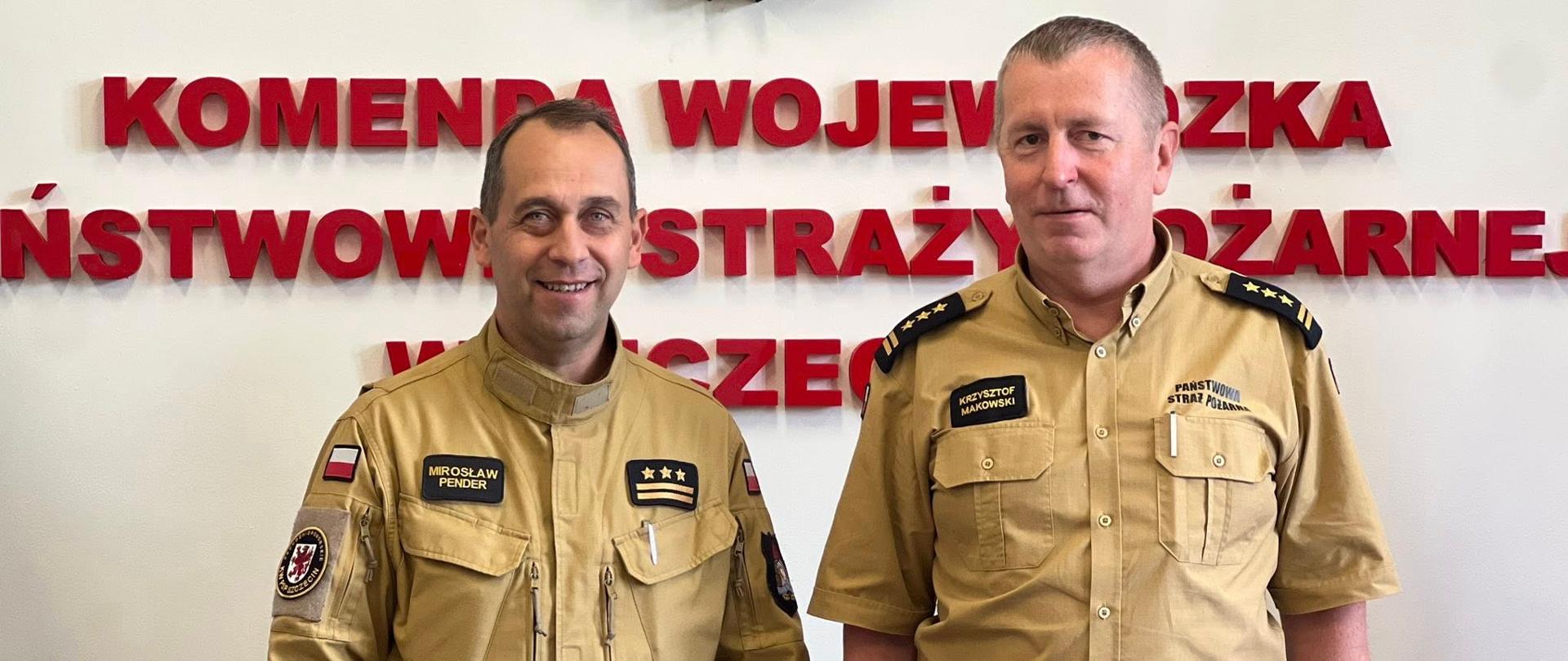 Na zdjęciu widać dwóch strażaków ubranych w mundury w kolorze piaskowym. Stoją obok siebie, uśmiechając się lekko. Za nimi na ścianie znajduje się duże godło Państwowej Straży Pożarnej – herb z polskim orłem w czerwonym tle, otoczony złotymi liśćmi oraz szarą hełmową oprawą z czerwonym płomieniem na górze. Pod orłem widnieje niebieska wstęga z napisem „PAŃSTWOWA STRAŻ POŻARNA”. Pod godłem znajdują się czerwone, trójwymiarowe litery z napisem w języku polskim: „KOMENDA WOJEWÓDZKA PAŃSTWOWEJ STRAŻY POŻARNEJ”. 