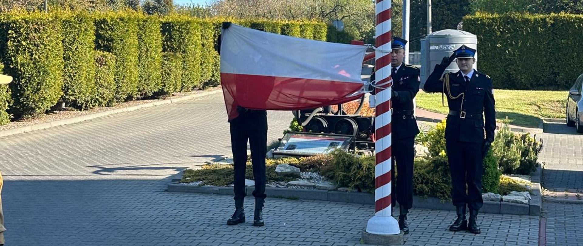 Na zdjęciu przedstawiono uroczystość podniesienia flagi państwowej na maszt. Funkcjonariusze Państwowej Straży Pożarnej w umundurowaniu galowym dokonują wciągnięcia flagi Rzeczypospolitej Polskiej, a jeden z nich oddaje honor.
Wydarzenie odbywa się na terenie jednostki, w otoczeniu uporządkowanej infrastruktury i zieleni. Uroczystość ma charakter oficjalny i związana jest z podkreśleniem szacunku dla symboli narodowych.