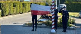 Na zdjęciu przedstawiono uroczystość podniesienia flagi państwowej na maszt. Funkcjonariusze Państwowej Straży Pożarnej w umundurowaniu galowym dokonują wciągnięcia flagi Rzeczypospolitej Polskiej, a jeden z nich oddaje honor.
Wydarzenie odbywa się na terenie jednostki, w otoczeniu uporządkowanej infrastruktury i zieleni. Uroczystość ma charakter oficjalny i związana jest z podkreśleniem szacunku dla symboli narodowych.