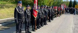 Widok z boku. Po lewej stronie pododdział strażaków OSP w Marcyporębie w mundurach wyjściowych. W tle drzewa. 