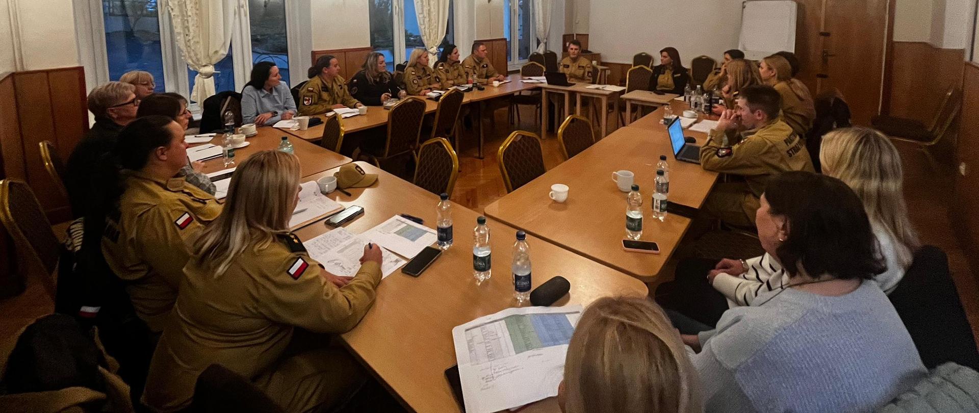 Zdjęcie przedstawia spotkanie strażaków siedzących przy dużych, złączonych stołach w sali konferencyjnej. Większość uczestników nosi piaskowe mundury, a część osób cywilne ubrania. Na stołach leżą dokumenty, notatki i butelki z wodą. Uczestnicy rozmawiają i słuchają osoby prowadzącej spotkanie, a atmosfera wygląda na roboczą i spokojną.