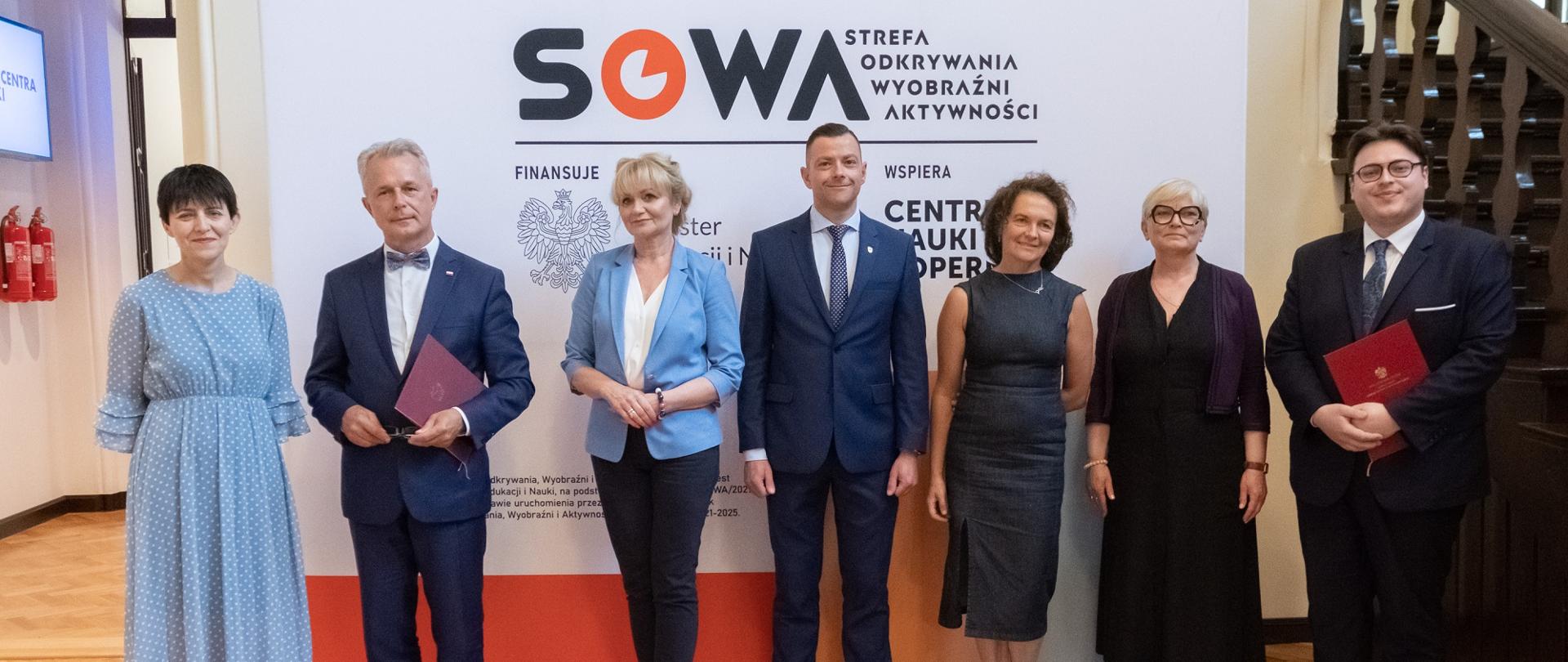 Uczestnicy uroczystości stoją na tle ścianki z napisem SOWA, obok korytarz i schody. fot. P.F. MATYSIAK