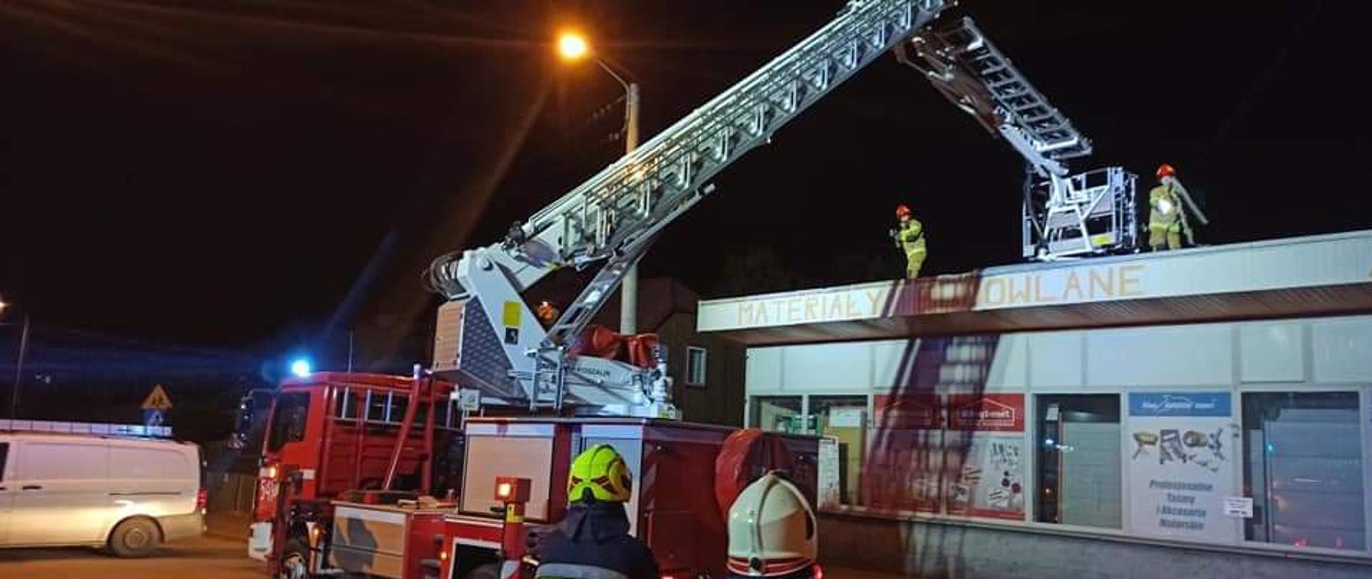 Na pierwszym planie strażacy w ubraniach specjalnych i hełmach ustawieni tyłem do fotografa. Za nimi znajduje się podnośnik hydrauliczny w trakcie działań ratowniczych. Obok podnośnika znajduje się budynek handlowy, z którego zerwało poszycie dachowe. Na dachu znajdują się kolejni strażacy, który zabezpieczają obiekt plandeką. Zdjęcie wykonane na drodze wieczorową porą.