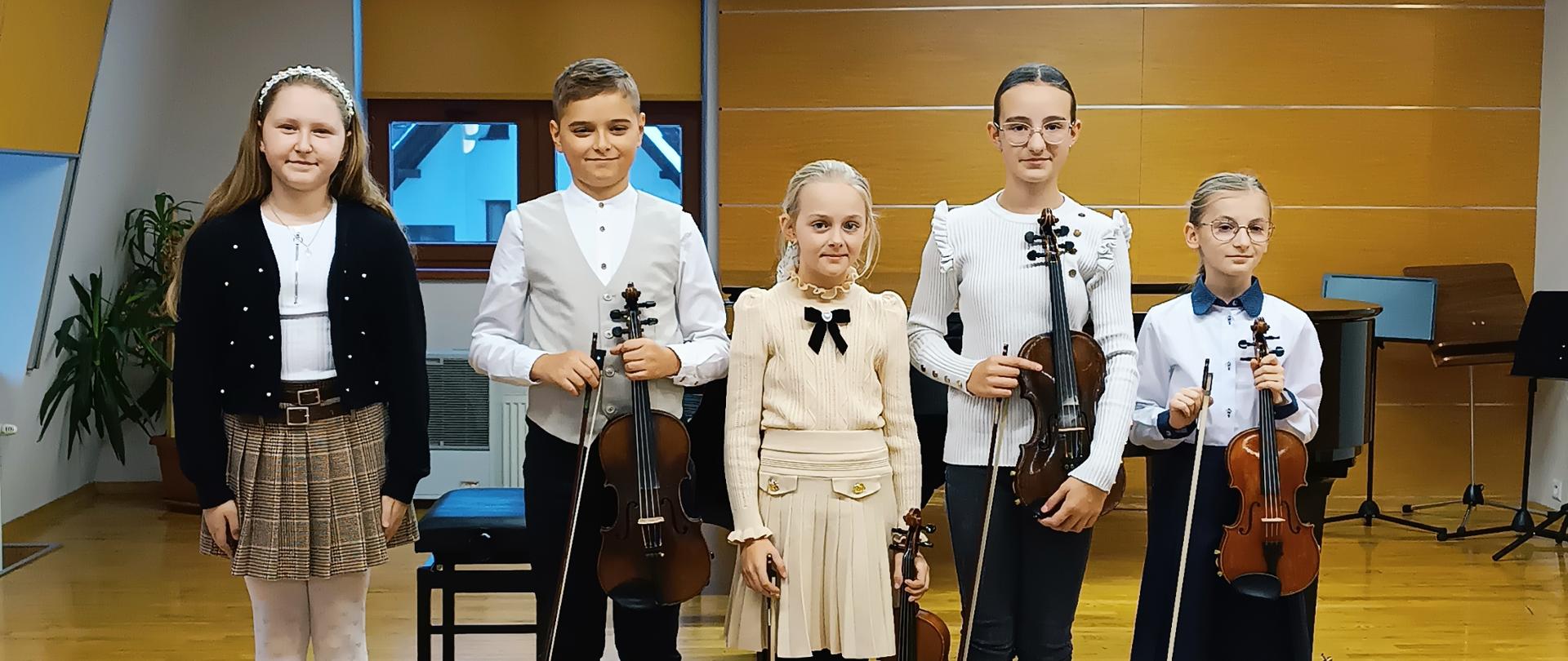 Na zdjęciu widać pięcioro dzieci stojących na scenie w jasnym, szkolnym lub muzycznym wnętrzu. Wszyscy są odświętnie ubrani i trzymają instrumenty – cztery osoby mają skrzypce, a jedna osoba (po lewej) nie ma instrumentu i wygląda, jakby mogła być uczestniczką występu albo częścią grupy, która jeszcze nie gra. Od lewej strony: Pierwsza dziewczynka ma długie włosy, opaskę, ciemny sweterek, jasną spódniczkę w kratkę i białe rajstopy. Obok stoi chłopiec w białej koszuli i czarnych spodniach, trzymający skrzypce w dłoni. W środku stoi dziewczynka w jasno¬beżowej sukience, również ze skrzypcami. Następnie dziewczynka w białej bluzce i ciemnych spodniach, trzymająca skrzypce i smyczek. Ostatnia, z prawej strony, to dziewczynka w białej koszuli i granatowej spódnicy, także ze skrzypcami. Wszyscy stoją równo obok siebie, uśmiechnięci, jak do pamiątkowego zdjęcia z koncertu lub zajęć muzycznych. Tło to jasna, drewniana ściana i podłoga sceny, z elementami sali szkolnej: oknem, rośliną i stojakami na nuty.