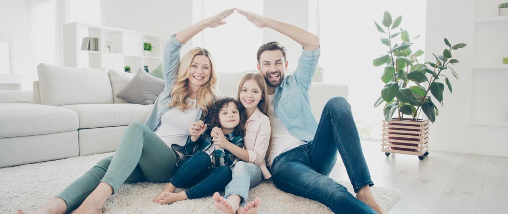 Czteroosobowa rodzina siedzi na białym dywanie, w tle jasne mieszkanie, kanapa i kwiat. Rodzice trzymają nad głowami dzieci ręce w górze tworząc wzór dachu domu