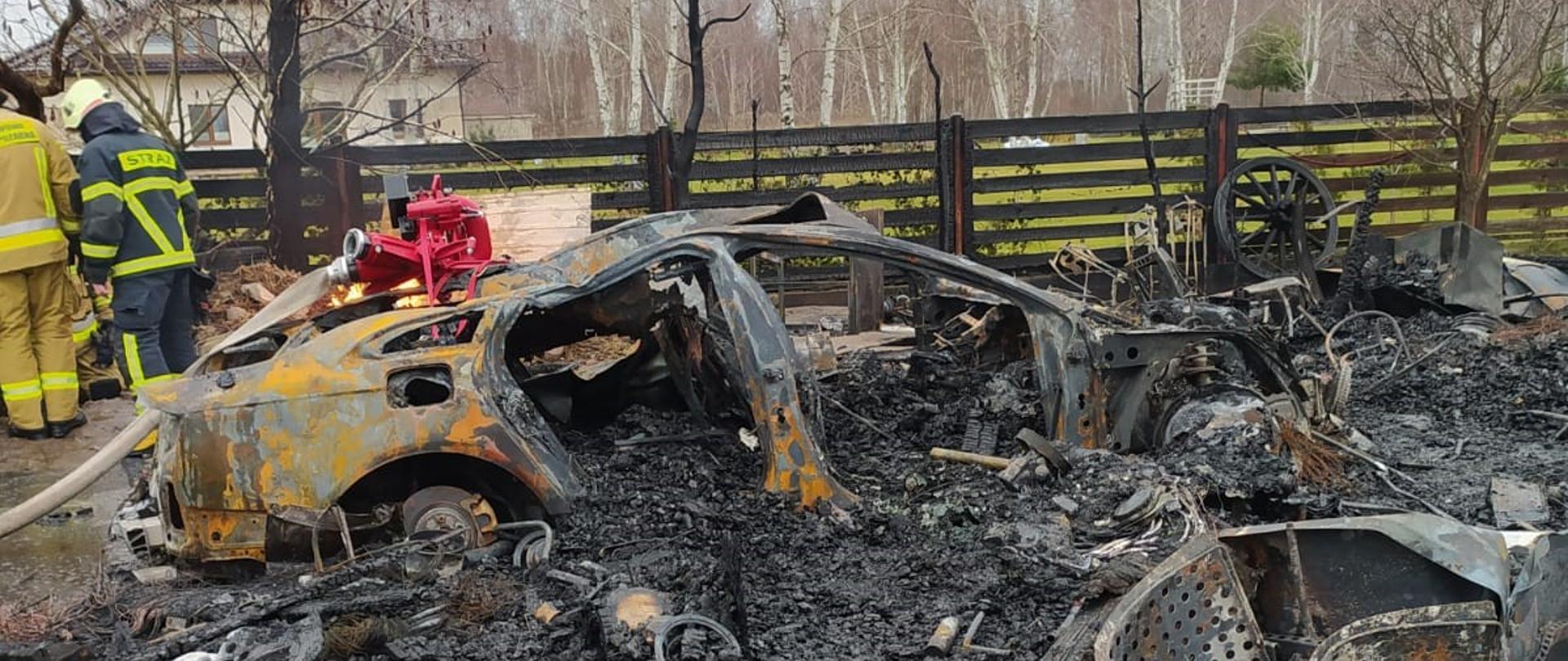 Na pierwszym planie widać zgliszcza po pożarze, w dalszym planie widac karoserię spalonego samochodu osobowego. 