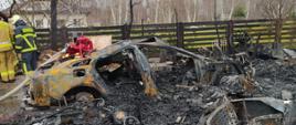 Na pierwszym planie widać zgliszcza po pożarze, w dalszym planie widac karoserię spalonego samochodu osobowego. 