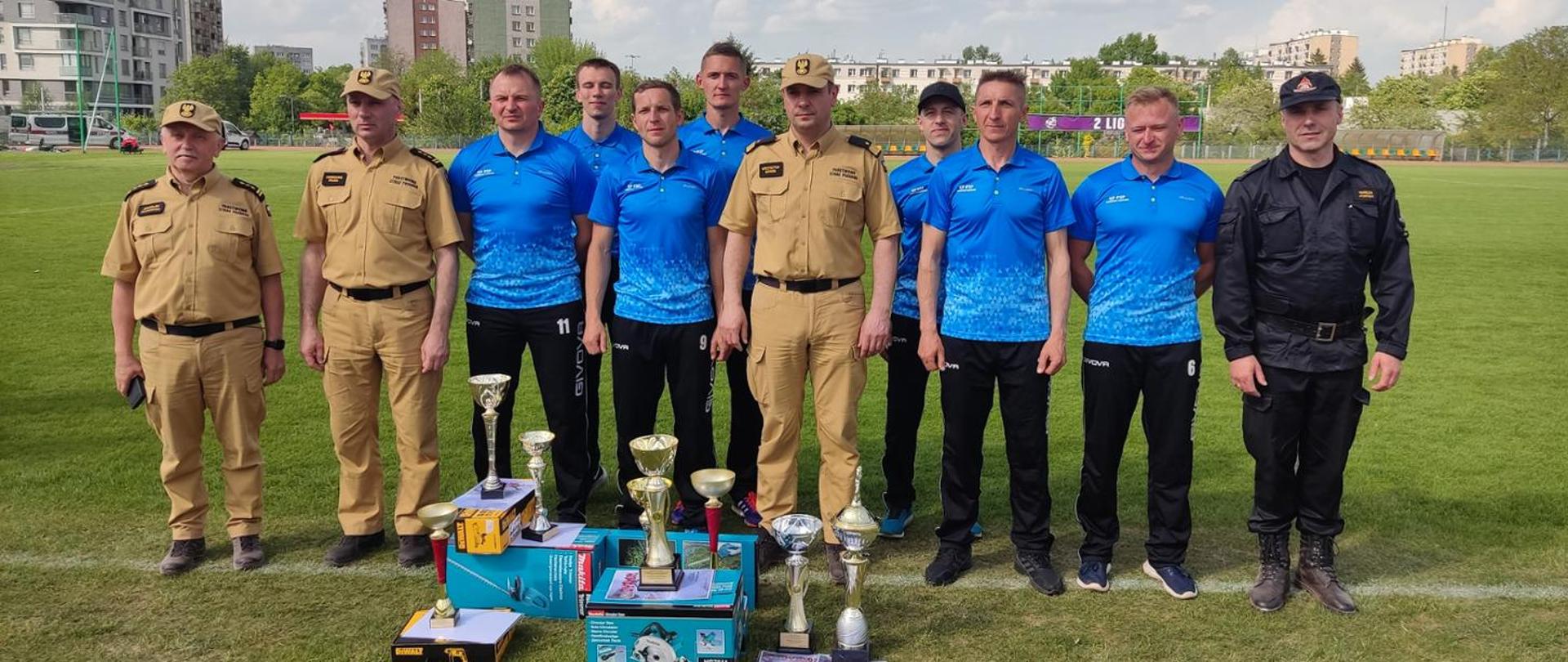 Zdjęcie na pierwszym planie przedstawia grupę zawodników ubranych w niebieskie koszulki i czarne spodnie. W grupie zawodników widać przedstawicieli KW PSP w Kielcach ubranych w ubrania służbowe koloru piaskowego. Przed grupą zawodników stoją ustawione puchary. Zawodnicy stoją na murawie stadionu. W oddali widać bloki mieszkalne.