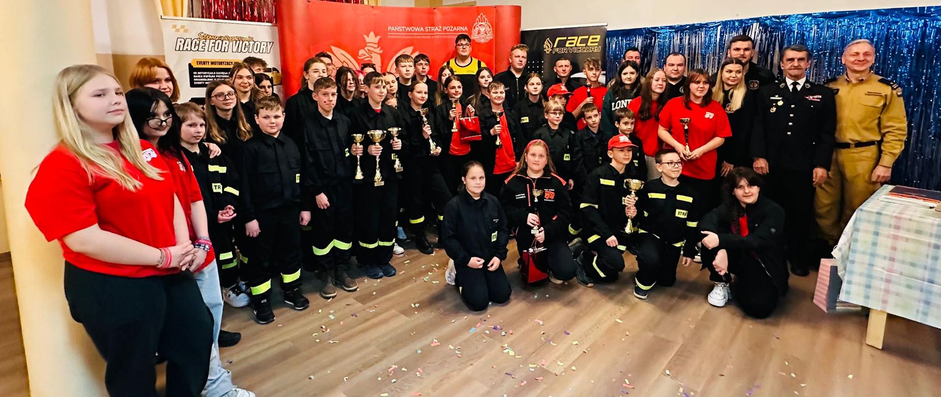 Grupa strażaków, młodzieży i dzieci pozuje do wspólnego zdjęcia w udekorowanej sali podczas turnieju UNO. Uczestnicy stoją i klęczą w kilku rzędach, część z nich trzyma puchary i nagrody. W tle widoczne są kolorowe girlandy, banery oraz dekoracje w barwach czerwonych i niebieskich, a na podłodze rozsypane jest konfetti. Atmosfera jest radosna i uroczysta.

