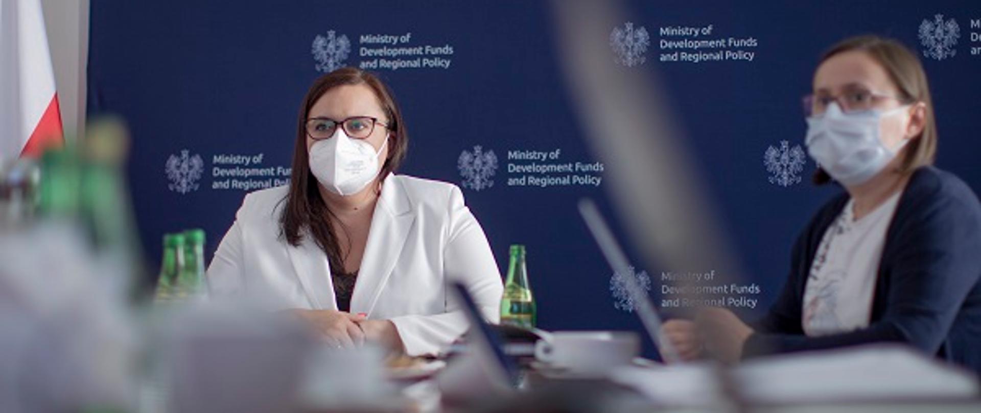 Wiceminister Małgorzata Jarosińska-Jedynak i pracownica MFiPR siedzą przy stole konferencyjnym. W tle ścianka z logo resortu w języku angielskim.