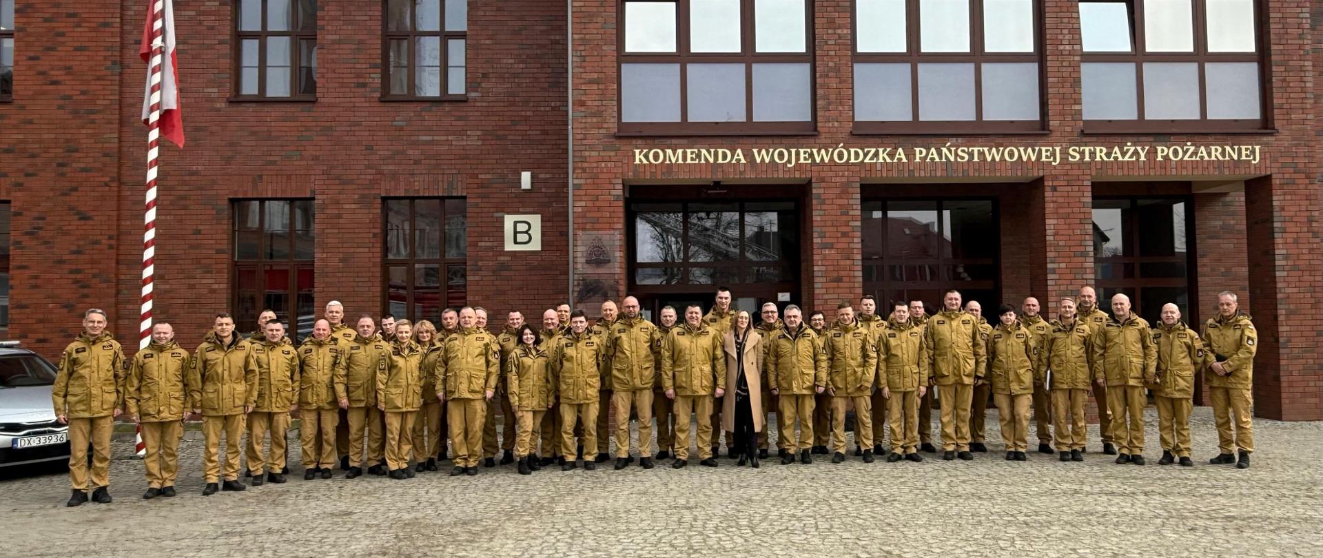  Zdjęcie grupowe strażaków Państwowej Straży Pożarnej na tle budynku Komendy Wojewódzkiej PSP. Wszyscy ubrani są w mundury i ustawieni w rzędach do oficjalnej fotografii.