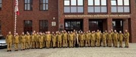 Zdjęcie grupowe strażaków Państwowej Straży Pożarnej na tle budynku Komendy Wojewódzkiej PSP. Wszyscy ubrani są w mundury i ustawieni w rzędach do oficjalnej fotografii.