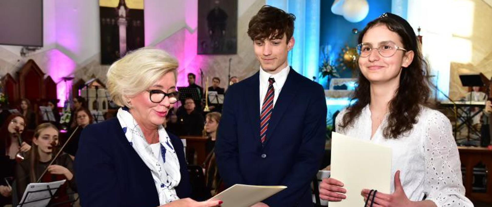 Minister Machałek czyta z kartki, za nią stoi dwoje elegancko ubranych uczniów.