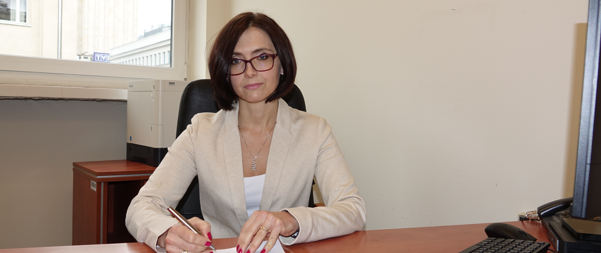 Kobieta siedząca za biurkiem, przy komputerze, podpisuje dokument