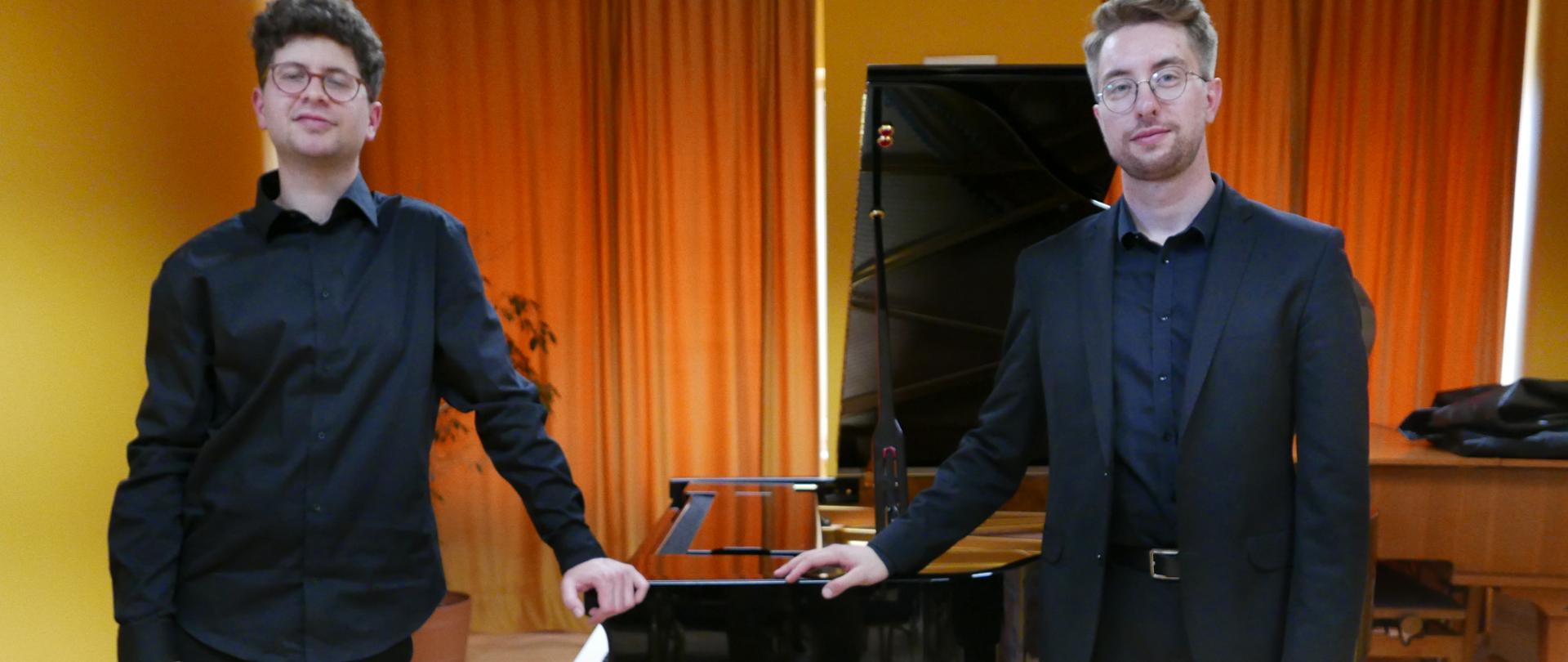 Na zdjęciu zrobionym w auli szkoły widzimy dwóch młodych pianistów pozujących do zdjęcia z fortepianem w tle. 