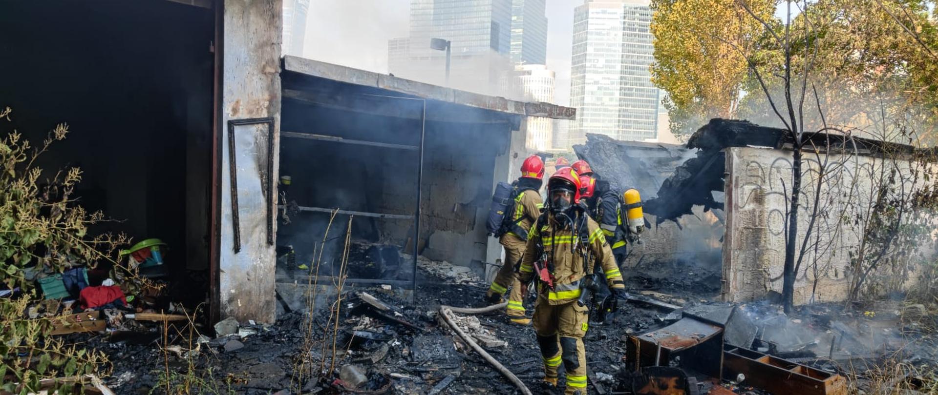 Na zdjęciu widać biały budynek pustostanu w którym doszło do pożaru oraz strażaków w piaskowych oraz granatowych ubraniach ochronnych. Na plecach ratowników widać butle od aparatów ochrony dróg oddechowych a na twarzach maski, które pomagają przebywać w strefie zadymieniu. Na głowach maja czerwone hełmy