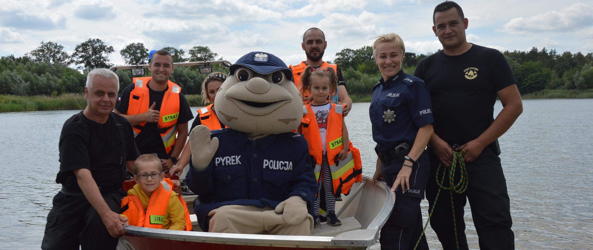 Kręci mnie bezpieczeństwo ... nad wodą. Akwen na ternie byłego poligonu wojskowego w Sierakowie. Na nim zwodowana jest strażacka łódź ratownicza. Na jej pokładzie policyjna maskotka Sierżant Pyrek, strażacy oraz mama dwójką dzieci. Obok łodzi znajdują się strażacy oraz policjantka. W tle drzewa Osoby znajdujące się w łódce mają ubrane kapoki. 